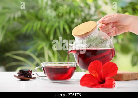 Frau, die köstlichen Hibiskus-Tee in eine Tasse am weißen Holztisch draußen gießt, Nahaufnahme Stockfoto