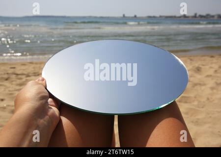 Frau mit rundem Spiegel, der den Himmel am Strand reflektiert, Nahaufnahme Stockfoto