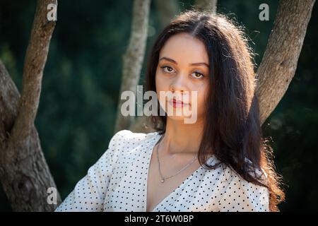 Eine junge Frau mit weißem und blauem Polka Dot-Shirt steht draußen vor einer Reihe von Bäumen und lächelt in die Kamera Stockfoto