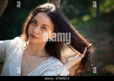 Eine schöne junge Frau mit langen, dunklen Haaren steht auf einer sonnendurchfluteten Wiese, ein Baum im Hintergrund Stockfoto