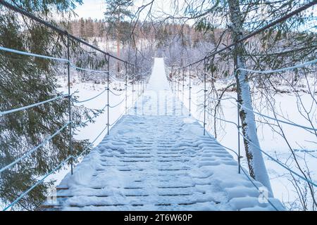 Wunderschöne, an einem Seil hängende Holzbrücke über den Fluss. Winterlandschaft, schneebedeckter Wald. Perspektive, Symmetrie Stockfoto