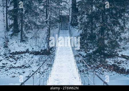 Wunderschöne, an einem Seil hängende Holzbrücke über den Fluss. Winterlandschaft, schneebedeckter Wald. Perspektive, Symmetrie Stockfoto