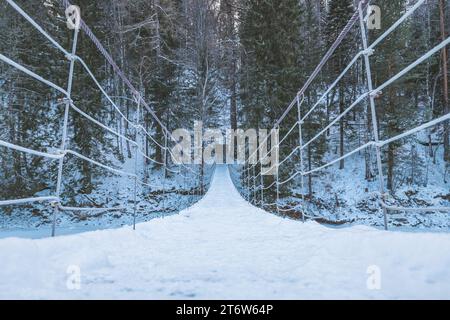 Wunderschöne, an einem Seil hängende Holzbrücke über den Fluss. Winterlandschaft, schneebedeckter Wald. Perspektive, Symmetrie Stockfoto