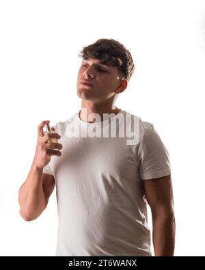 Ein Mann in einem weißen Hemd, der eine Parfümflasche hält und sie auf seinen Hals spritzt. Schöner Mann in einem weißen Hemd, der Köln sprüht Stockfoto