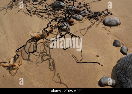 Gebrochene Stücke von Wire Gabion Basket Flood Defences am Strand, der von hochenergetischen Wellen aus Schindeln und Schattenmustern auf dem Sand entfernt wurde. Stockfoto