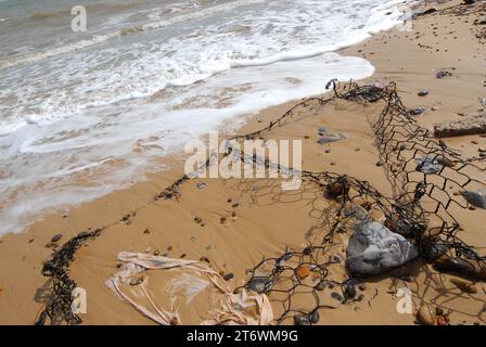 Schwarze Bruchstücke von Gabion Wire Basket Flood Defences erzeugen Schattenmuster im Sand am Strand, der von hochenergetischen Wellen aus Schindeln befreit wird. Stockfoto