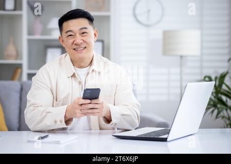 Ein glücklicher Koreaner sitzt in einem hellen Wohnzimmer an einem Tisch vor einem Laptop, hält ein Telefon in der Hand, tippt auf den Bildschirm, schreibt, lächelt und sieht in die Kamera. Stockfoto