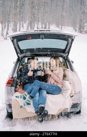 Frau lacht mit einer Tasse, die neben dem Mann sitzt und Kaffee trinkt und den Daumen im Kofferraum hochhält Stockfoto
