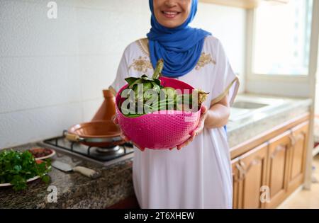Details auf einer rosafarbenen Schüssel mit gewaschenem frischem Bio-Gemüse in den Händen einer Hausfrau, die einen blauen Hijab und ein authentisches weißes marokkanisches Kleid trägt, stan Stockfoto
