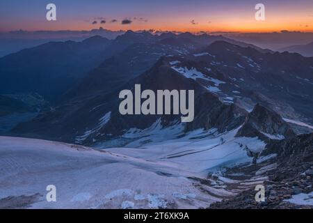 Alpiner Sonnenaufgang über kleinem Gletscher in den italienischen Alpen. Mehrere Berggipfel kurz vor Sonnenaufgang über dem Horizont. Stockfoto