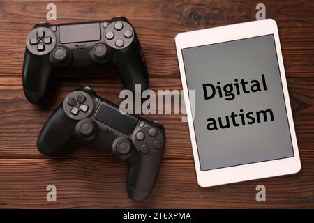Tablet mit Phrase Digital Autism und Gamepads auf Holztisch, flach gelegt. Suchtverhalten Stockfoto
