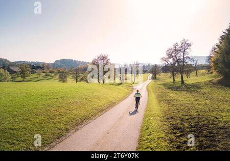 Weibliche Radfahrerin, die auf einer Landstraße in der Nähe von grünen Wiesen und bunten Wäldern fährt, genießt die herbstliche Landschaft an einem sonnigen Tag, Luftaufnahme. Stockfoto