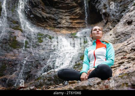 Junge Frau, die Yoga praktiziert, sitzt in eigener Pose auf dem Felsen in der Nähe des wunderschönen Wasserfalls. Entspannungskonzepte, Natur und Wohlbefinden. Stockfoto