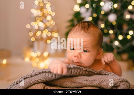 Silvester Weihnachtsfeierszene - lustiger süßer neugeborener Junge, der auf einer gestrickten Decke liegt, mit weihnachtlichen Weihnachtsbaumspielzeugen und Lichtergirlande. Kinderartikel-Konzept, Kinderzimmer Stockfoto