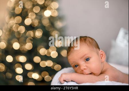 Silvester Weihnachtsfeierszene - lustiger süßer neugeborener Junge, der auf einer gestrickten Decke liegt, mit weihnachtlichen Weihnachtsbaumspielzeugen und Lichtergirlande. Kinderartikel-Konzept, Kinderzimmer Stockfoto