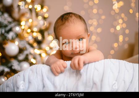 Silvester Weihnachtsfeierszene - lustiger süßer neugeborener Junge, der auf einer gestrickten Decke liegt, mit weihnachtlichen Weihnachtsbaumspielzeugen und Lichtergirlande. Kinderartikel-Konzept, Kinderzimmer Stockfoto