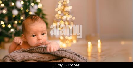 Silvester Weihnachtsfeierszene - lustiger süßer neugeborener Junge, der auf einer gestrickten Decke liegt, mit weihnachtlichen Weihnachtsbaumspielzeugen und Lichtergirlande. Kinderartikel-Konzept, Kinderzimmer Stockfoto