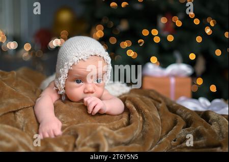 Silvester Weihnachtsfeierszene - niedlicher neugeborener Junge in lustiger Mütze auf gestrickter beiger Decke mit weihnachtlichem Baum Gold Spielzeug und Lichtergirlande. Kinderartikel-Konzept, Kinderzimmer Stockfoto