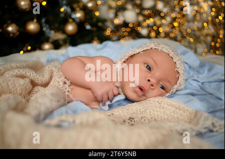 Silvester Weihnachtsfeierszene - niedlicher neugeborener Junge in lustiger Mütze auf gestrickter beiger Decke mit weihnachtlichem Baum Gold Spielzeug und Lichtergirlande. Kinderartikel-Konzept, Kinderzimmer Stockfoto