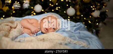 Silvester Weihnachtsfeierszene - niedlicher neugeborener Junge in lustiger Mütze auf gestrickter beiger Decke mit weihnachtlichem Baum Gold Spielzeug und Lichtergirlande. Kinderartikel-Konzept, Kinderzimmer Stockfoto