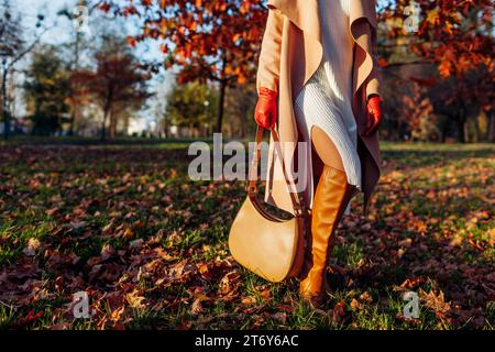 Damen-Outfit im Herbst. Modische Frau mit stilvollem weißem Strickkleid, kniehohen Stiefeln, beigefarbenem Mantel, Handschuhen mit Handtasche im Park unter gefallenem Urlaub Stockfoto