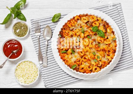 Tortellini gebacken mit Mozzarella-Käse, Tomatensoße und Rinderhack in weißer Backform auf weißem Holztisch, horizontale Sicht von oben, flach liegend Stockfoto