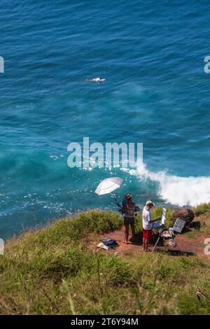 Zwei Personen plein Air Painting auf einer Klippe mit Blick auf das türkisblaue tropische Wasser, während ein Surfer auf Maui, Hawaii, USA, vorbeipaddelt. Stockfoto