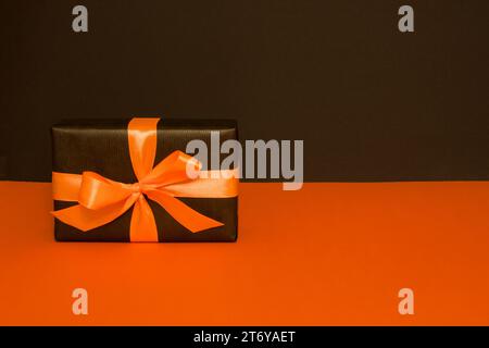 Eine rechteckige Geschenkbox aus schwarzem Papier, verziert mit einem orangefarbenen Satinband, auf schwarzem und orangefarbenem Hintergrund Stockfoto