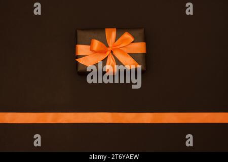 Eine rechteckige Geschenkbox ist in schwarzem Papier verpackt, verziert mit orangefarbenem Satinband auf schwarzem Hintergrund. Flache Lagen Stockfoto
