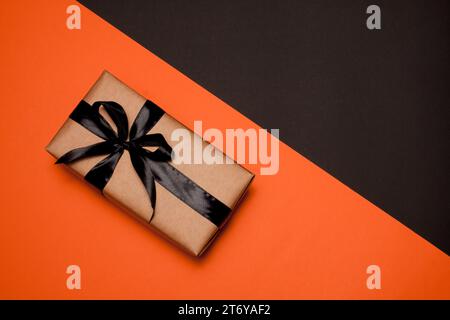 Eine rechteckige Geschenkbox, die in Kraftpapier gewickelt ist und mit einem schwarzen Satinband und einer Schleife verziert ist, liegt auf einem orange-schwarzen Hintergrund. Verkauf. Black Friday. Stockfoto