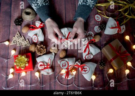 Adventskalender zu Weihnachten. Frau, die einen Beutel mit Geschenk in der Hand hält. Taschen mit Geschenken, Walnüssen, Konen, Soutache, Band, Boxen, Girlande, Zahlen, Wald Stockfoto