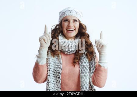 Hallo Winter. Lächelnde, stilvolle Frau in Pullover, Fäustlingen, Mütze und Schal vor weißem Hintergrund, der auf dem Kopierraum nach oben zeigt. Stockfoto