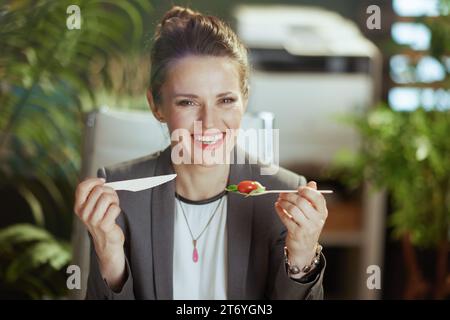 Nachhaltiger Arbeitsplatz. Glückliche, moderne 40-jährige Geschäftsfrau in einem grauen Business-Anzug im modernen grünen Büro, die Salat isst. Stockfoto