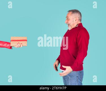 Überraschter reifer Mann, der die Hand der Frau sieht, die eine Geschenkbox vor türkisfarbenem Hintergrund hält Stockfoto