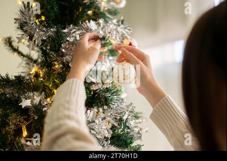 Nahaufnahme einer Frau, die einen Weihnachtsbaum mit einem schönen Ornament dekoriert und eine Weihnachtskugel an einen Weihnachtsbaum hängt. Spezielles Urlaubskonzept Stockfoto