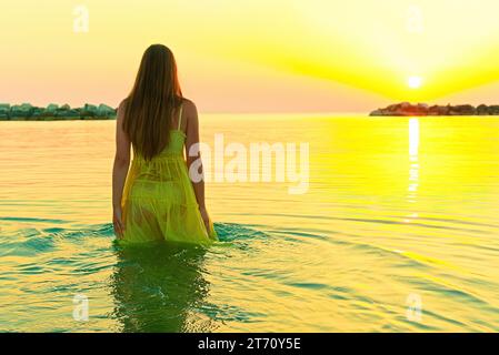 Eine junge Frau in einem hellgelben Kleid begrüßt den Sonnenaufgang, indem sie ins Meer geht. Stockfoto