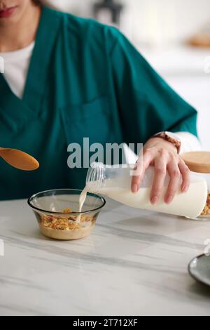 Zugeschnittenes Bild einer medizinischen Krankenschwester, die nicht-Milchmilch in Schüssel mit Müsli hinzufügte Stockfoto