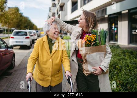 Reife Enkelin mit Großmutters Einkaufstasche. Seniorin und Betreuerin, die im kalten Herbst mit Lebensmitteln aus dem Supermarkt nach Hause gehen Stockfoto