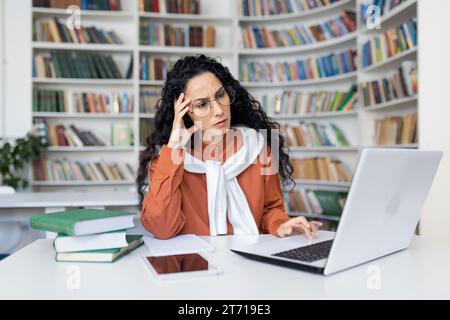 Müde junge hispanische Lehrerin sitzt am Schreibtisch vor dem Laptop im Schulzimmer und hält Hand auf Kopf, Überarbeit im Büro Kopfschmerzen, Online-Lernkonzept. Stockfoto