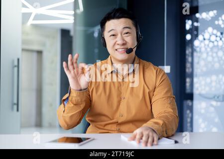 Glücklicher asiatischer Büroangestellter, der im hellen Büro am Schreibtisch sitzt und ein Headset trägt und bei Videogesprächen spricht, ein chinesischer Geschäftsmann lächelt und ein gutes Schild zeigt, der in die Kamera blickt und die Kamera betrachtet. Stockfoto