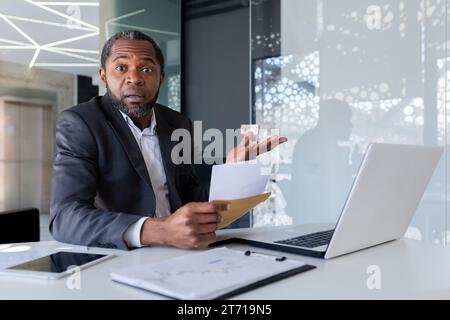 Ein Erwachsener überraschte den offenen Umschlag in den Händen, schockiert von Nachrichten, enttäuschenden Nachrichten, afroamerikanischer Chef sitzt im hellen Büro und blickt in die Kamera und spreizt die Hände. Stockfoto