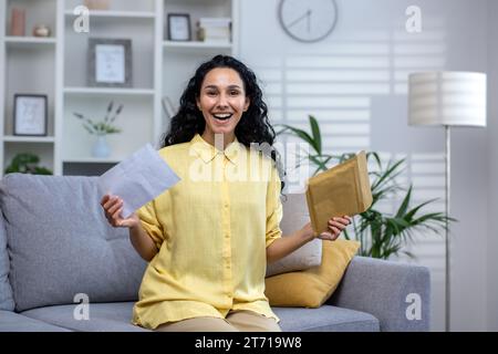Glückliche, lockige hispanische Frau, die zu Hause auf dem Sofa im Wohnzimmer sitzt und einen offenen Umschlag hält. Die süße Brünette ist glücklich mit den Nachrichten, gewinnt Lotterie, glückliche Nachrichten, lächelt und blickt in die Kamera. Stockfoto