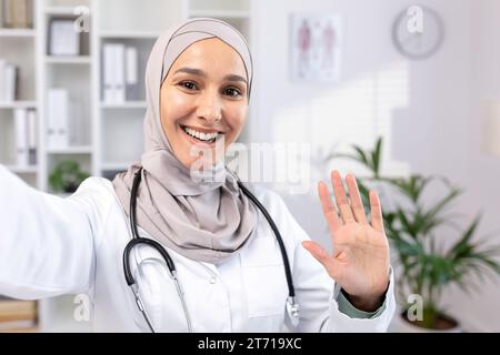 Schöne asiatische muslimische Ärztin in Hijab und Anzug, die Selfie-Fotos macht, während sie sich ausruhen. Ein Arzt führt einen Videoanruf durch, lächelt und winkt, während er seine Familie von seinem Arbeitsplatz aus begrüßt Stockfoto