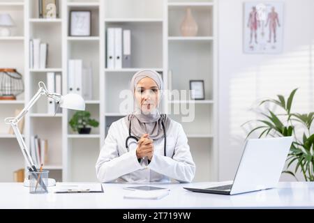 Porträt einer ernsthaften asiatischen muslimischen Ärztin im Hijab, die am Tisch im Krankenhausbüro mit Stethoskop um den Hals sitzt, Arme gefaltet, in die Kamera schauend. Stockfoto
