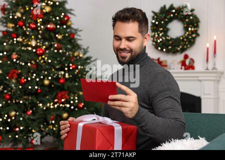 Glücklicher junger Mann mit Weihnachtsgeschenk Lesekarte zu Hause Stockfoto