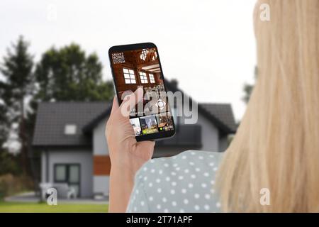 Frau, die ein intelligentes Heimsicherheitssystem auf dem Handy in der Nähe des Hauses im Freien benutzt. Gerät, das verschiedene Räume durch Kameras anzeigt Stockfoto