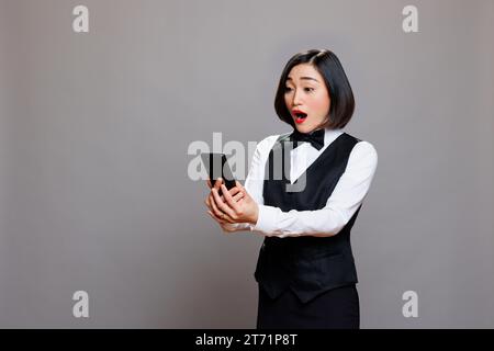 Schockiert zufriedene Empfangsdame, die die Kundenzufriedenheitsnachricht auf dem Smartphone überprüft. Aufgeregt junge asiatische Kellnerin in Schwarz-weiß-Uniform, die die positive Bewertung des Kunden auf dem Handy liest Stockfoto