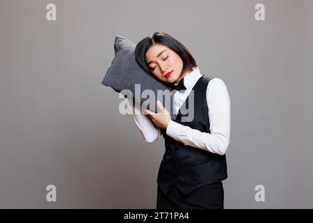 Abgenutzt attraktive asiatische Kellnerin in Restaurantuniform, die auf Kissen schläft. Verschlafene erschöpfte junge Empfangsdame mit geschlossenen Augen posiert im Studio auf grauem Hintergrund Stockfoto