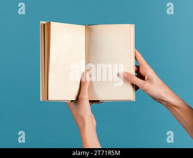 Anonyme schlanke junge Frau mit gepflegten Fingern, die leere Seiten gebundenes Buch in Licht halten und vor blauem Hintergrund demonstrieren Stockfoto