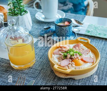 Frühstück mit Spiegeleiern in einer Pfanne mit Speck und Gemüse, Frühstück in einem Luxushotel in Thailand, gebratene Eier in einer kleinen Pfanne mit Schweinehackfleisch und Würstchen Stockfoto